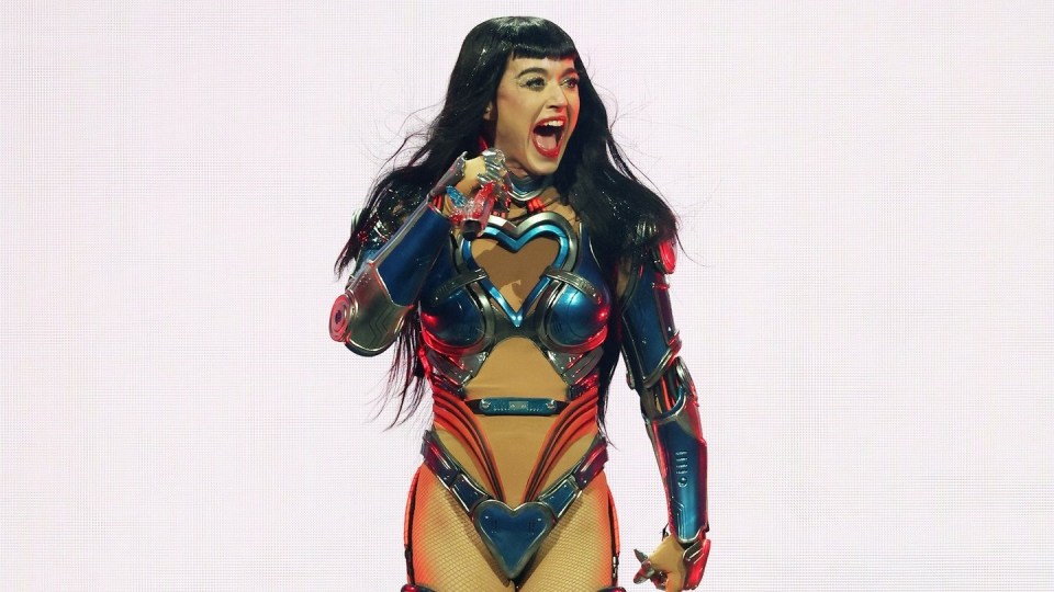 Woman with long black hair, wearing a blue and silver cut-out metal outfit, performing on-stage holding a microphone.