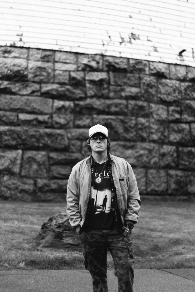 Black and white image of a man standing in front of a brick wall, wearing combat trousers, a jacket and a cap.