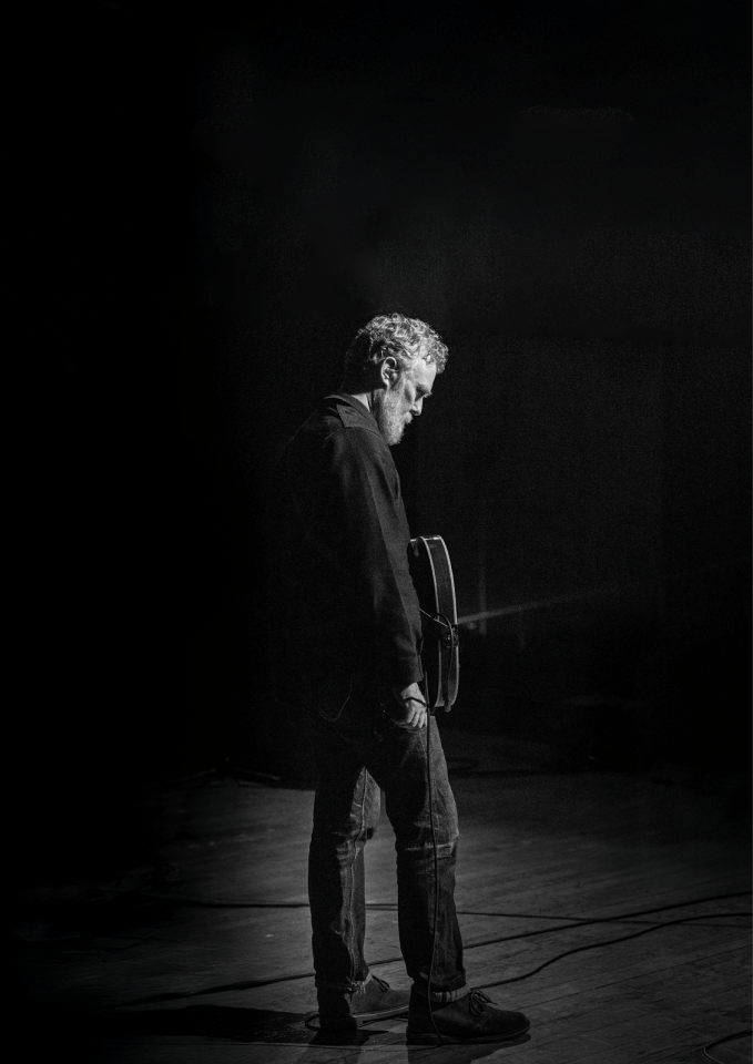 Black and white image of Glen Hansard holding a guitar, wearing all black, looking downwards at the ground.