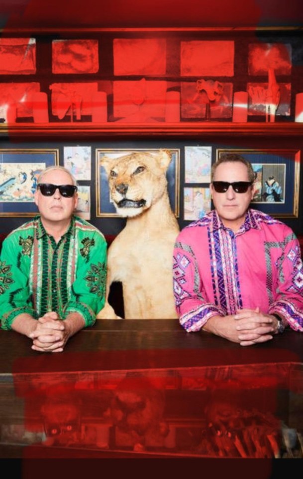 Two middle-aged men in colourful patterned shirts sitting on either side of a fake leopard at a red desk with a red background.