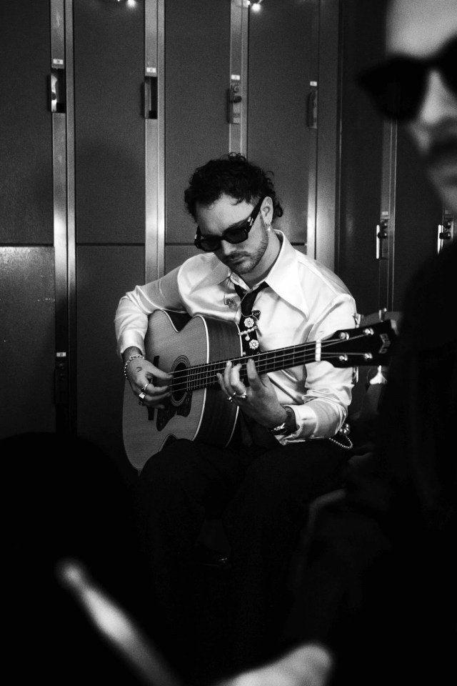 A young man wearing black sun glasses and a white shirt and tie, sitting down playing on his guitar