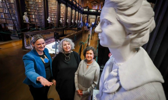 Three women in an old library looking at a white marble sculpture of a woman.